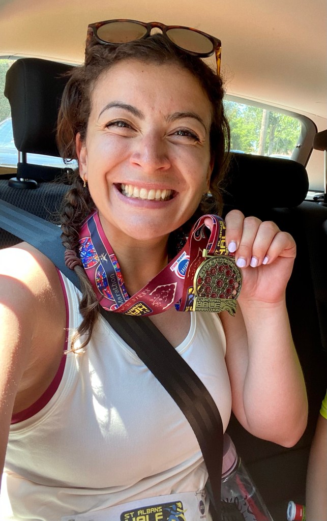 A woman sitting in a car smiling and holding a red and gold medal from the St Albans Half Marathon