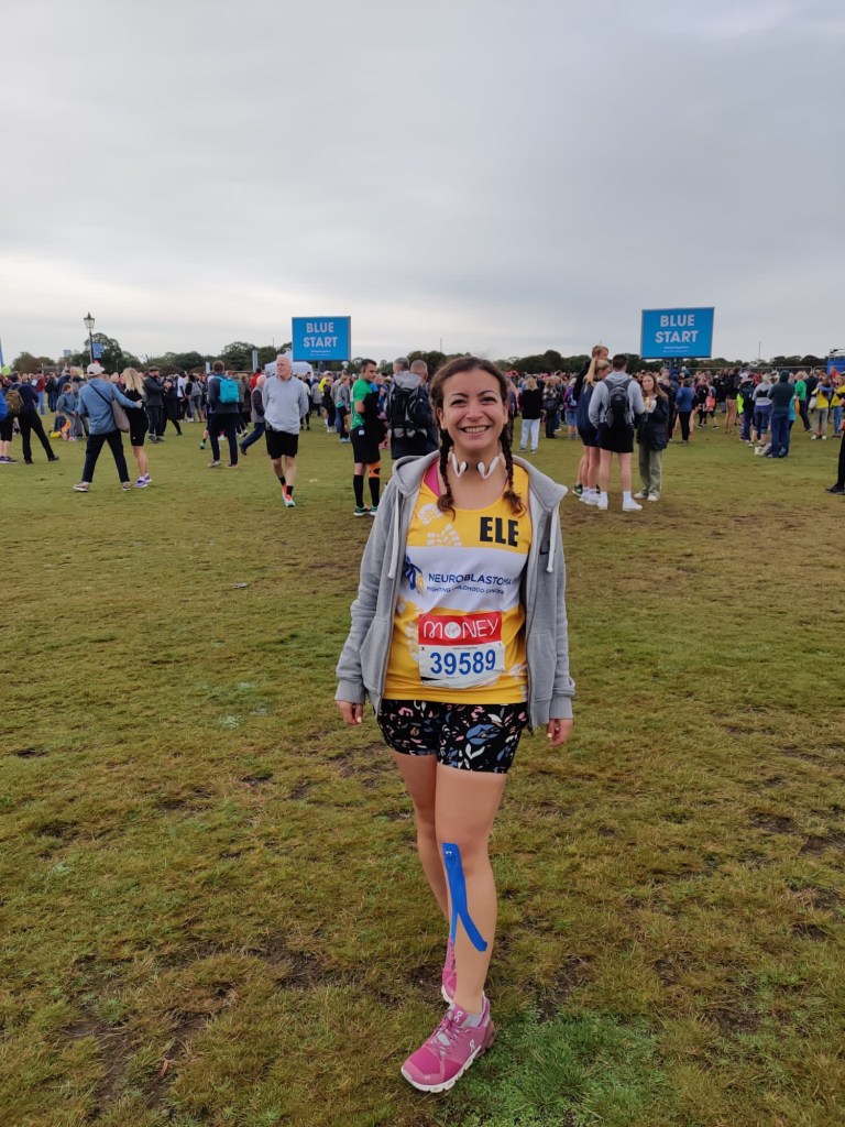 A woman standing in a field in Blackheath, London before the start of the London Marathon wearing shorts, a yellow vest and a grey jumper