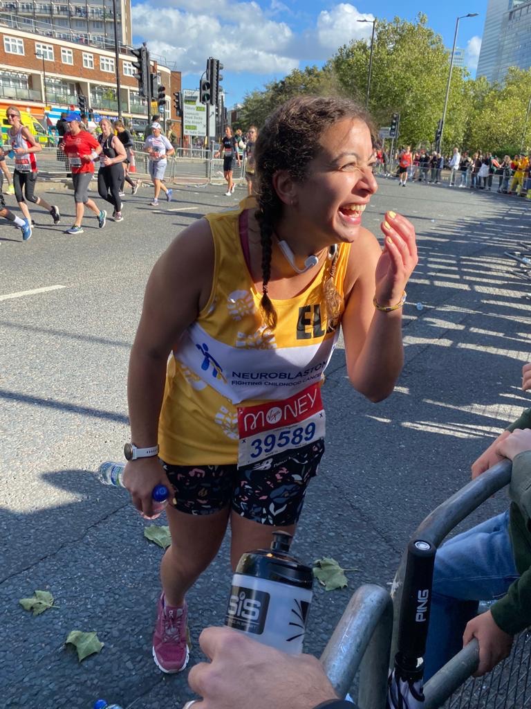 A woman laughing at the crowd at the London Marathon, wearing a yellow running vest and shorts
