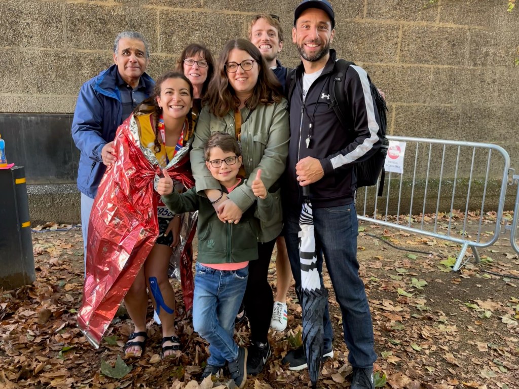 A family of seven people standing together to smile at the camera, with one wearing a red foil blanket after running a marathon