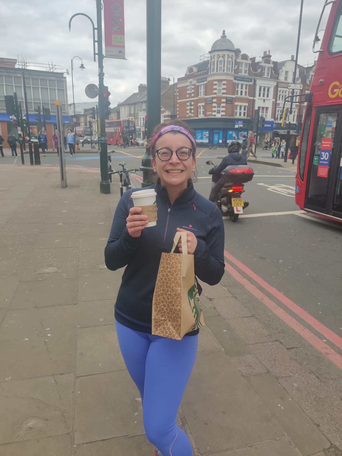 A woman standing on a pavement in Tooting, London holding a Starbucks coffee cup and carrier bag smiling at the camera