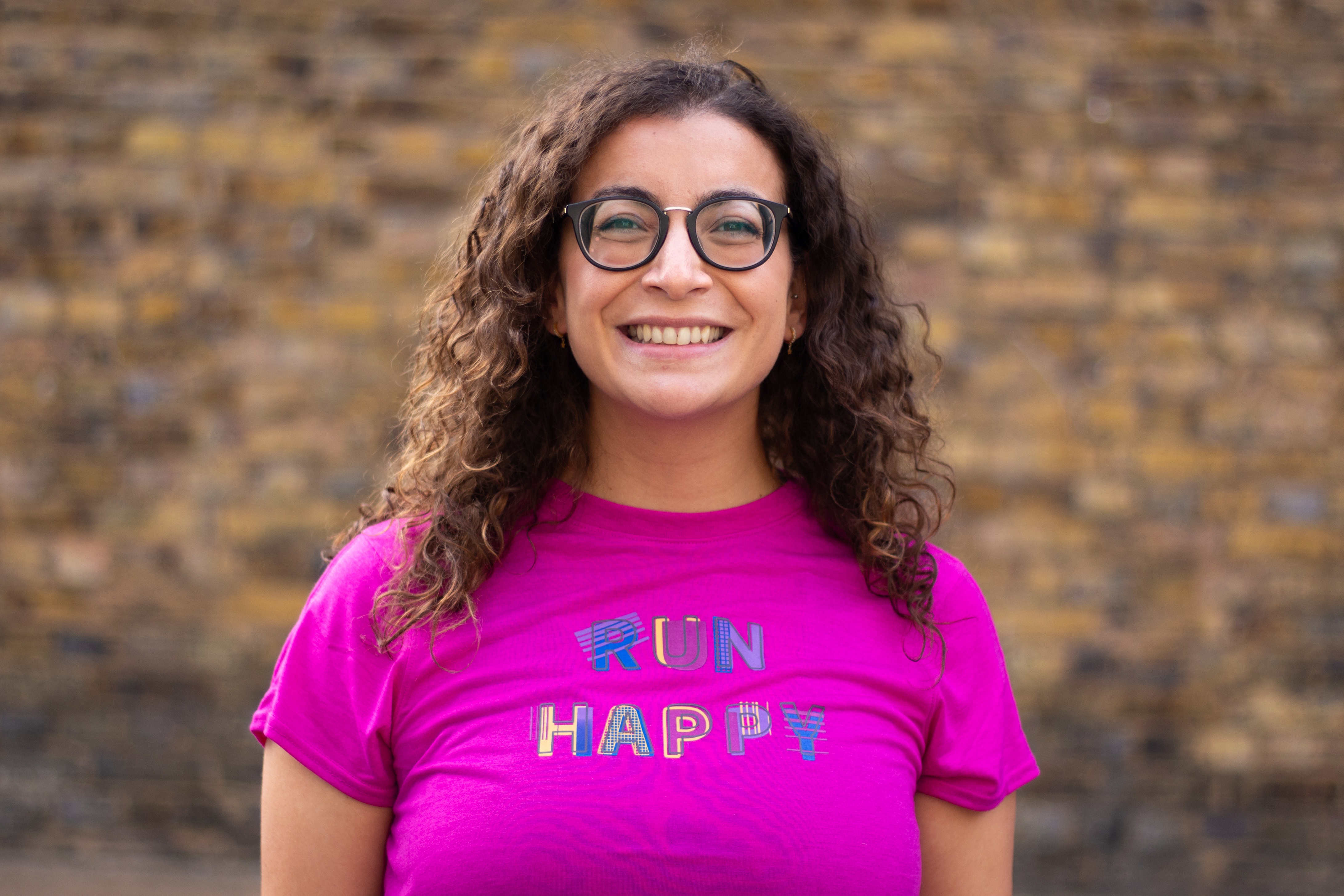A woman with brown curly hair and glasses smiling at the camera wearing a pink tshirt reading 'Run Happy'