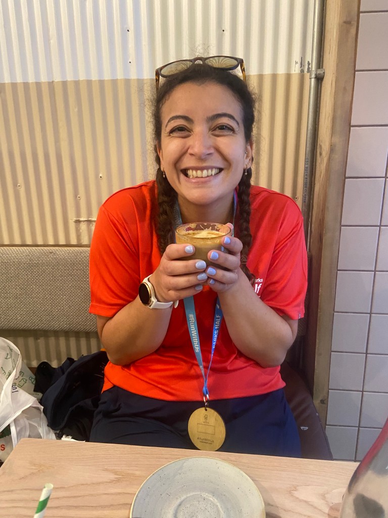 A woman in a red running t-shirt wearing a wooden medal from the Royal Parks Half Marathon smiling and holding a cup of coffee