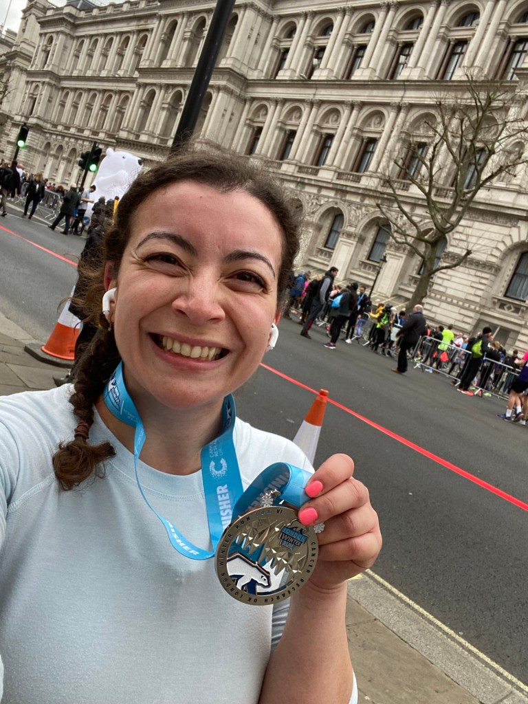A woman standing on the pavement in London wearing a blue running shirt and holding a medal from a 10k run