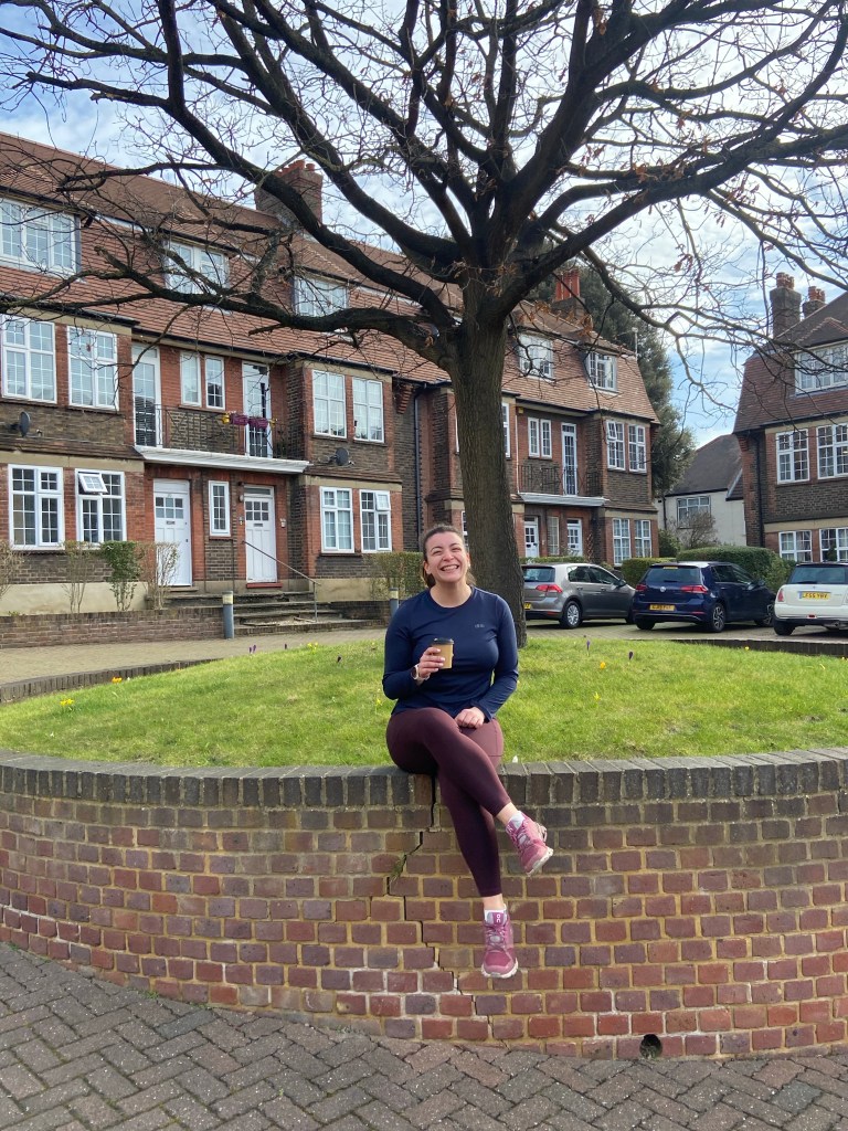 A woman sitting on a brick wall outside a building holding a coffee and wearing running clothes