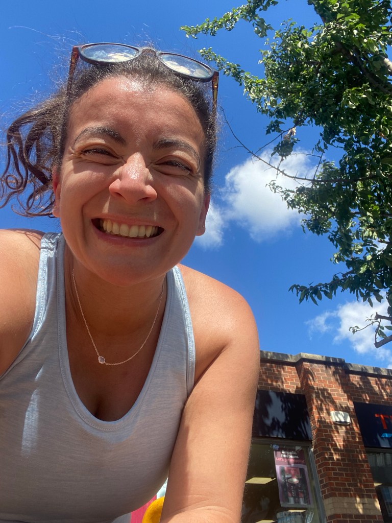 A woman smiling in a blue running vest with a bright blue sky and tree behind her