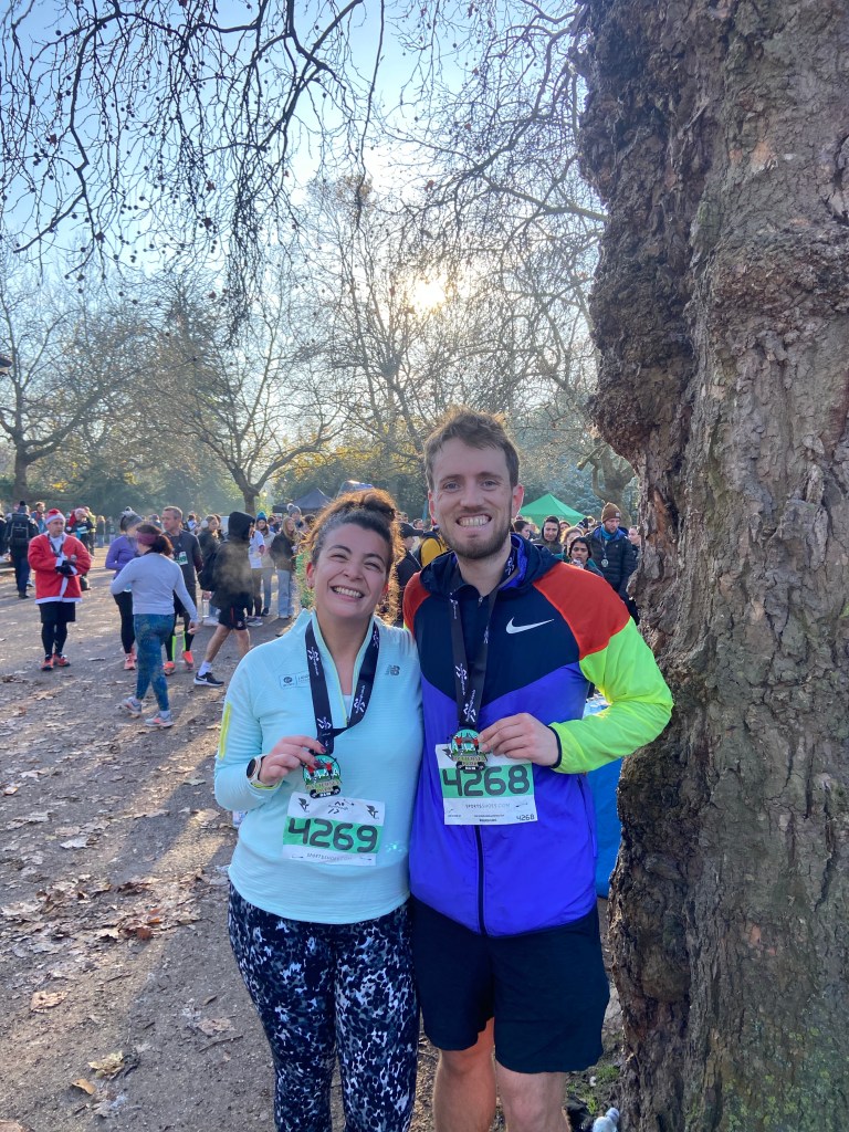 A man and a woman standing together in Battersea Park by a tree holding medals 