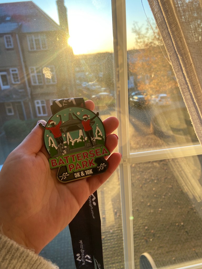 Medal from Battersea Park 5k & 10k race held in front of a window with the sun shining through