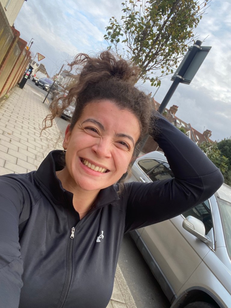 Image of a woman with brown curly hair in a ponytail and a black running t-short on smiling at the camera