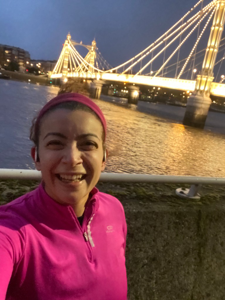 Selfie of a woman in a pink running jumper smiling in front of Albert Bridge in London with its lights on
