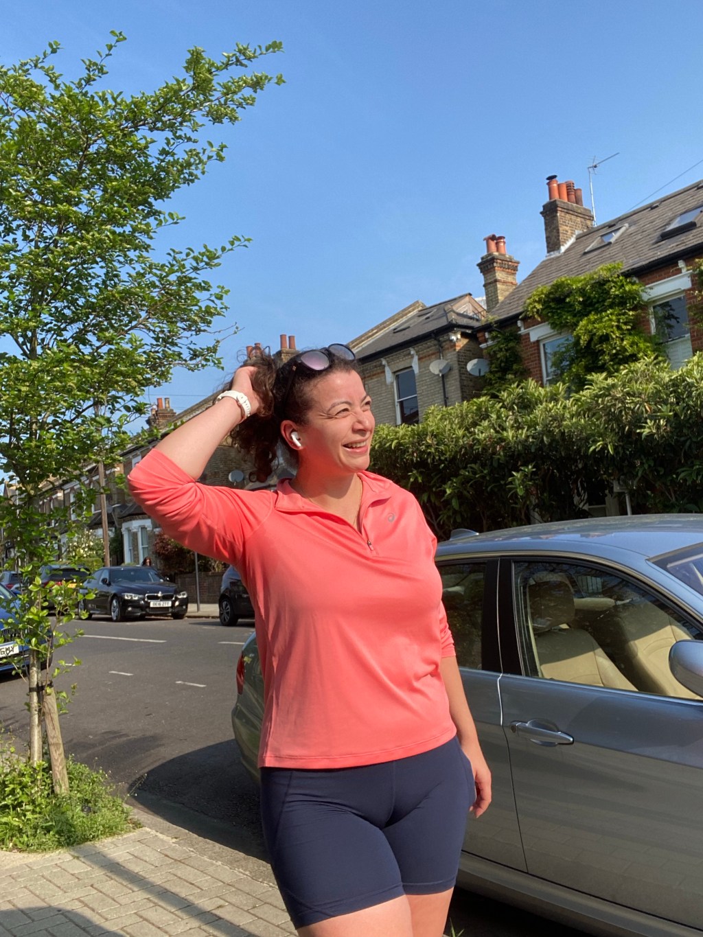 Photo of a woman with brown hair in a ponytail and sunglasses wearing blue running shorts and an orange running top standing on a pavement alongside houses