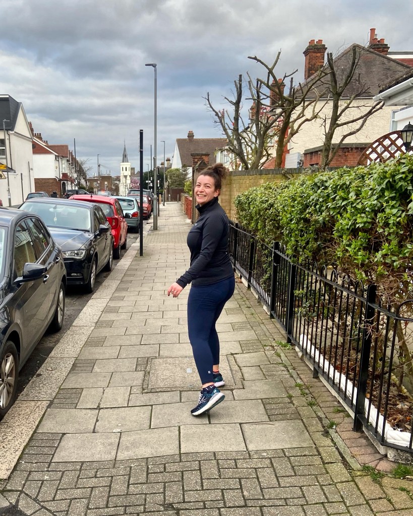 A girl on the pavement during a run smiling back at the camera in blue leggings, a black jumper and blue trainers
