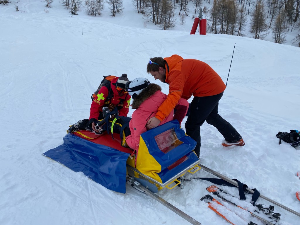 A girl on a skiing holiday being helped into a rescue stretcher on a snowy ski slope by two men